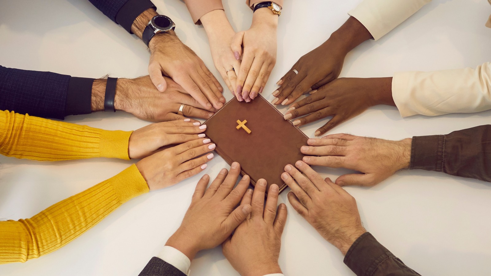Hands from many people reaching across a table to touch the tips of their fingers to a Bible.